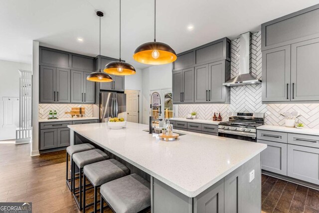 kitchen featuring appliances with stainless steel finishes, pendant lighting, wall chimney exhaust hood, and a kitchen island with sink