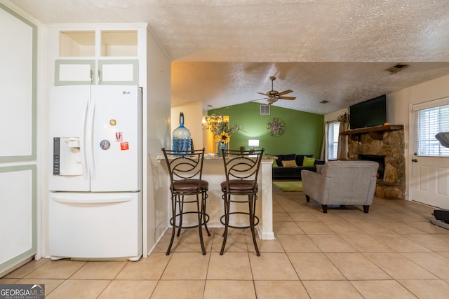 kitchen with a kitchen breakfast bar, light tile patterned floors, white fridge with ice dispenser, a stone fireplace, and vaulted ceiling