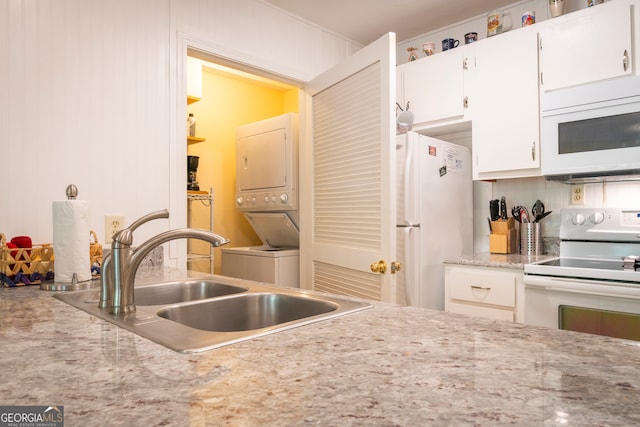 kitchen featuring stacked washing maching and dryer, white appliances, sink, light stone counters, and white cabinetry