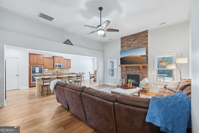 living room with a high ceiling, ceiling fan, crown molding, light hardwood / wood-style flooring, and a stone fireplace