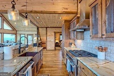 kitchen with light wood-type flooring, light stone counters, wall chimney range hood, hanging light fixtures, and stainless steel range