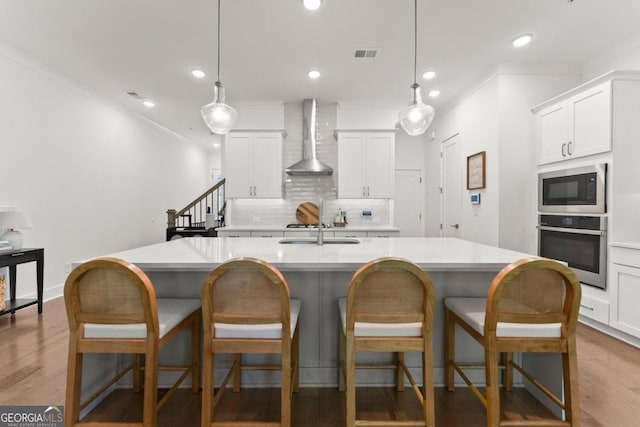 kitchen featuring wall chimney exhaust hood, a kitchen island with sink, decorative light fixtures, and stainless steel appliances