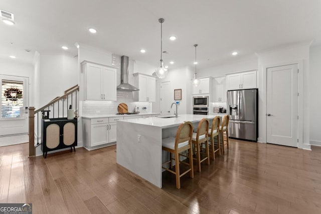 kitchen with appliances with stainless steel finishes, wall chimney range hood, a kitchen island with sink, and white cabinets