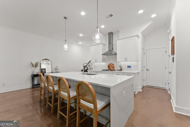 kitchen with white cabinets, wall chimney exhaust hood, light hardwood / wood-style floors, a center island with sink, and decorative light fixtures