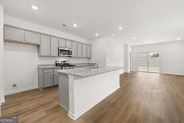 kitchen with light stone counters, gray cabinetry, a center island with sink, light hardwood / wood-style flooring, and appliances with stainless steel finishes