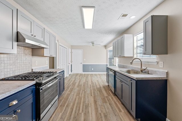 kitchen with a textured ceiling, appliances with stainless steel finishes, a sink, light wood finished floors, and under cabinet range hood