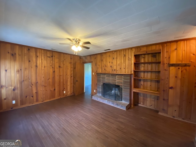 unfurnished living room with a brick fireplace, wooden walls, built in shelves, and dark wood-type flooring