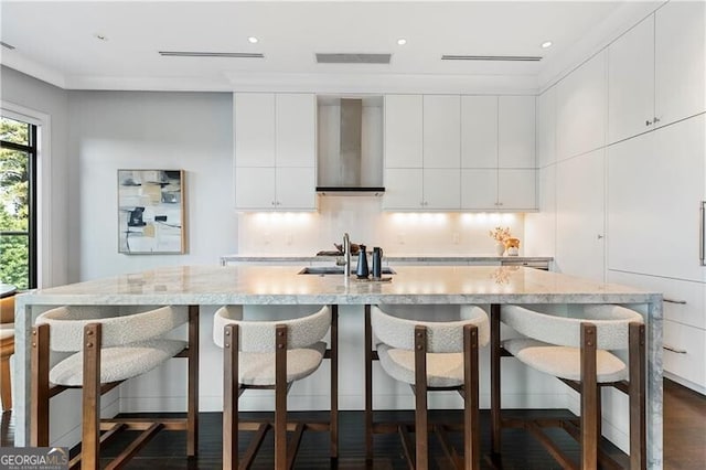 kitchen with wall chimney exhaust hood, dark hardwood / wood-style flooring, a large island with sink, sink, and white cabinets