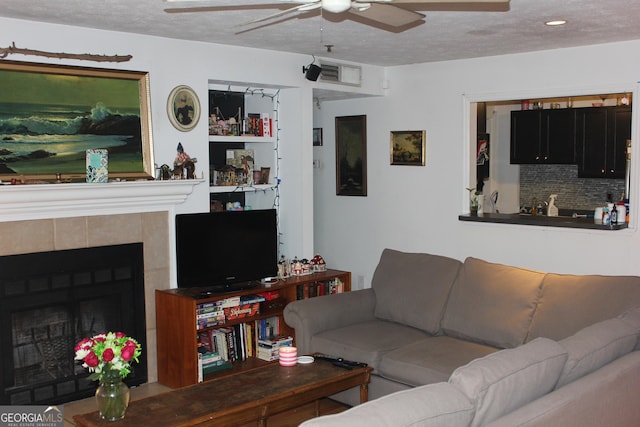 living room featuring ceiling fan, a textured ceiling, and a tiled fireplace