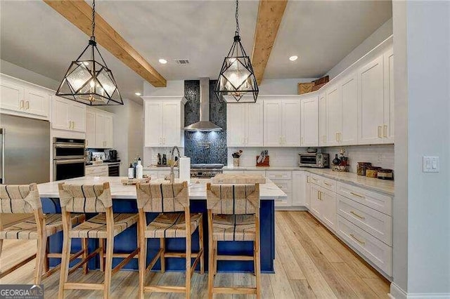 kitchen with beam ceiling, white cabinetry, a kitchen island with sink, and wall chimney range hood