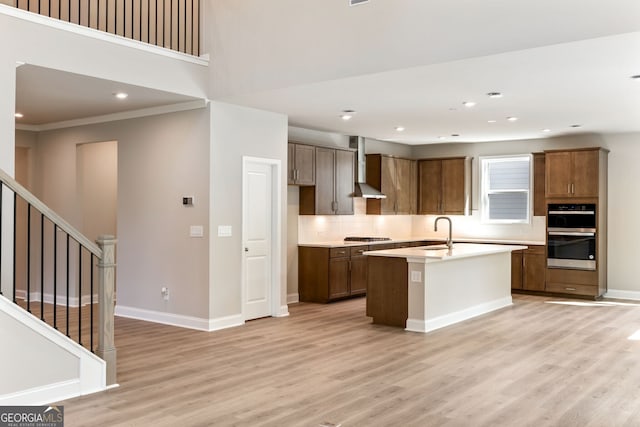 kitchen featuring an island with sink, ornamental molding, light hardwood / wood-style flooring, wall chimney exhaust hood, and appliances with stainless steel finishes