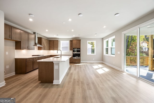 kitchen featuring stainless steel appliances, wall chimney range hood, light hardwood / wood-style floors, and a kitchen island with sink