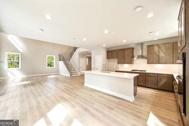 kitchen with wall chimney range hood, light wood-type flooring, a center island with sink, sink, and stainless steel appliances