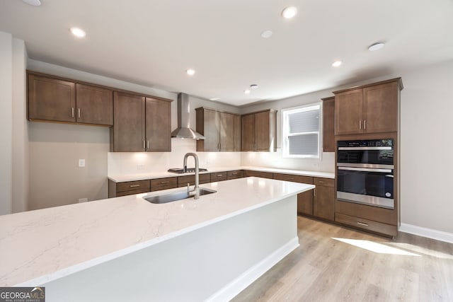 kitchen with light stone countertops, stainless steel double oven, sink, light hardwood / wood-style floors, and wall chimney range hood