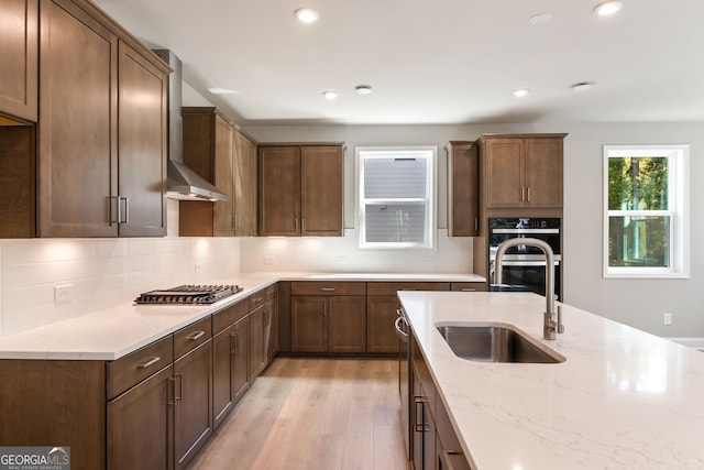 kitchen with light stone counters, stainless steel appliances, wall chimney exhaust hood, sink, and light hardwood / wood-style flooring