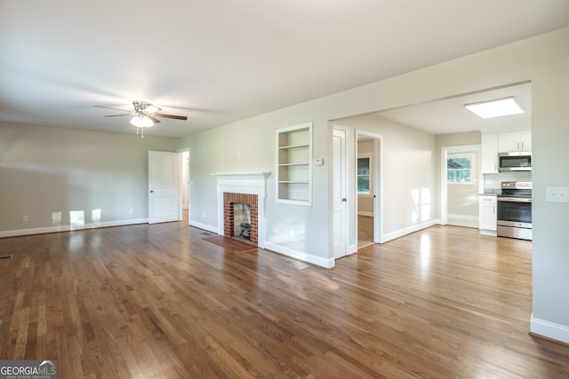 unfurnished living room featuring dark wood-type flooring, a brick fireplace, built in shelves, and ceiling fan