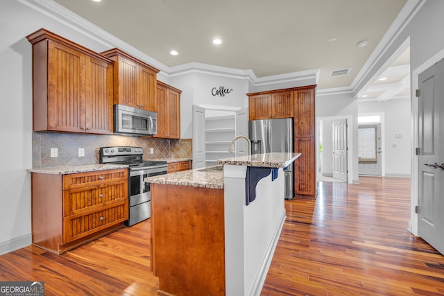 kitchen featuring an island with sink, light stone counters, light hardwood / wood-style floors, crown molding, and appliances with stainless steel finishes