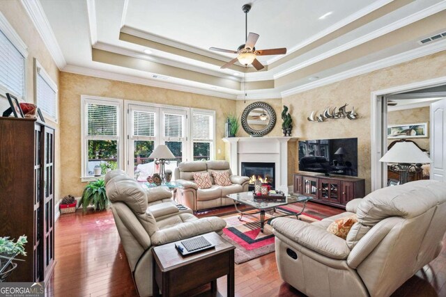 living room with dark wood-type flooring, ceiling fan, ornamental molding, and a tray ceiling