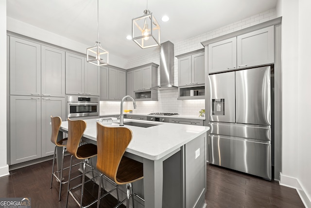 kitchen featuring wall chimney range hood, appliances with stainless steel finishes, a kitchen island with sink, sink, and decorative light fixtures