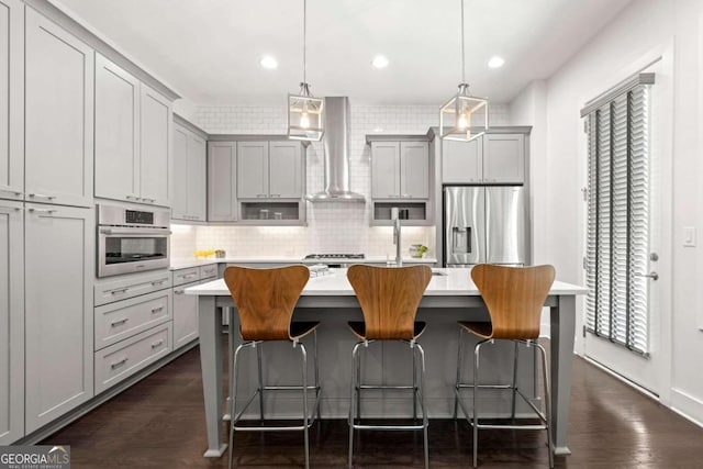 kitchen with wall chimney range hood, an island with sink, stainless steel appliances, pendant lighting, and gray cabinets