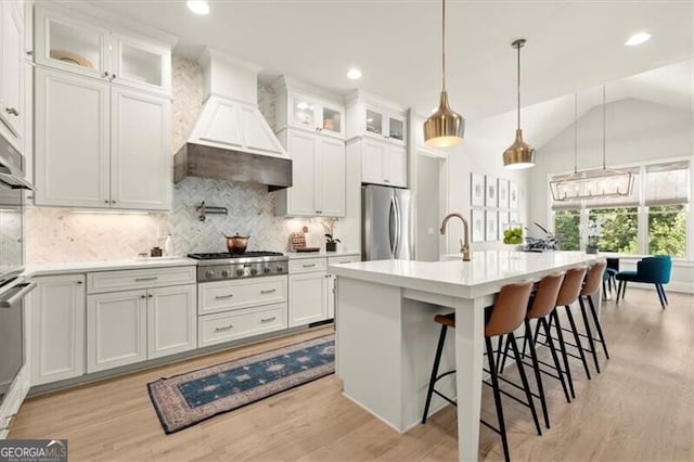 kitchen with an island with sink, hanging light fixtures, custom range hood, stainless steel appliances, and vaulted ceiling