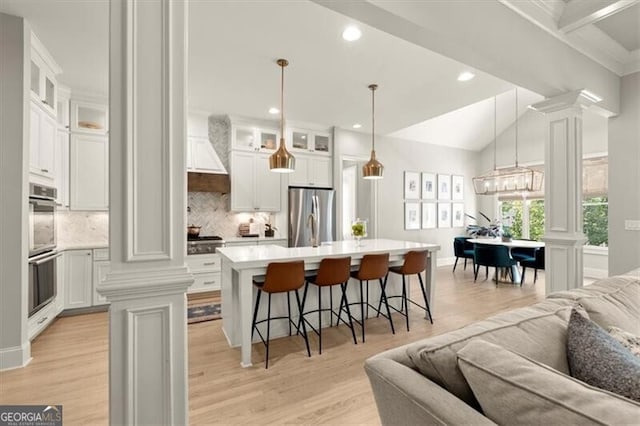 kitchen featuring light wood-type flooring, ornate columns, stainless steel appliances, white cabinets, and a kitchen island with sink