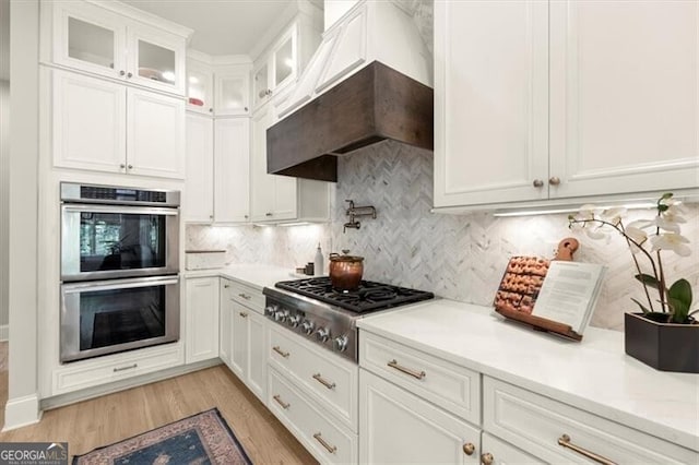 kitchen with custom exhaust hood, decorative backsplash, white cabinets, light wood-type flooring, and stainless steel appliances