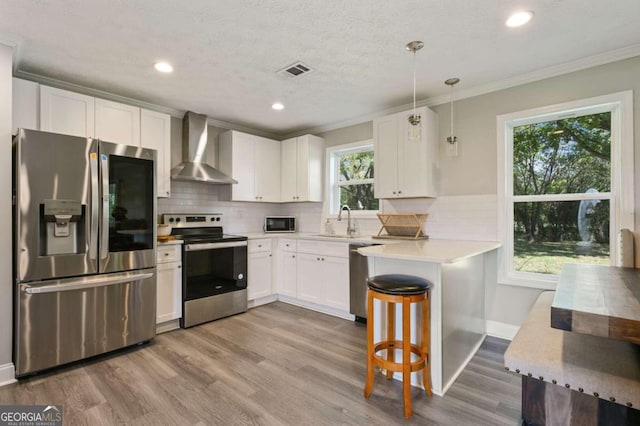 kitchen featuring wall chimney range hood, stainless steel appliances, a healthy amount of sunlight, decorative light fixtures, and white cabinetry