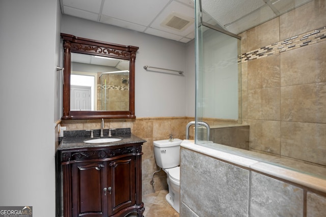 bathroom featuring toilet, a shower with shower door, vanity, a drop ceiling, and tile walls