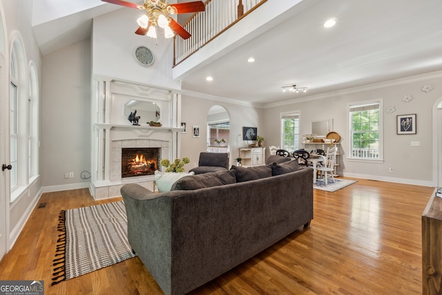 living room featuring a tiled fireplace, ceiling fan, high vaulted ceiling, light wood-type flooring, and ornamental molding