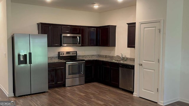 kitchen featuring sink, dark brown cabinets, stainless steel appliances, dark wood-type flooring, and light stone counters