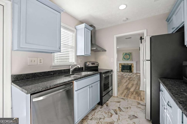 kitchen featuring a stone fireplace, wall chimney range hood, stainless steel appliances, dark stone counters, and sink