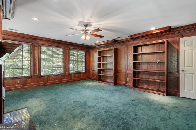 unfurnished living room with ceiling fan, wooden walls, dark colored carpet, ornamental molding, and built in shelves