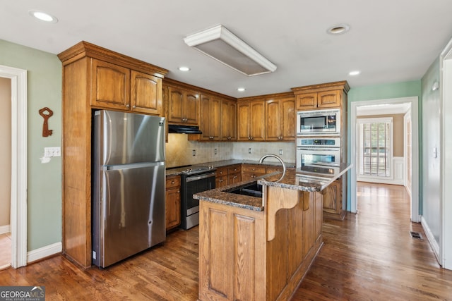 kitchen featuring dark wood-type flooring, stainless steel appliances, a center island with sink, and dark stone counters