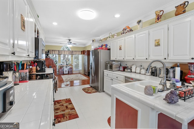 kitchen with appliances with stainless steel finishes, ceiling fan, tile counters, white cabinets, and crown molding