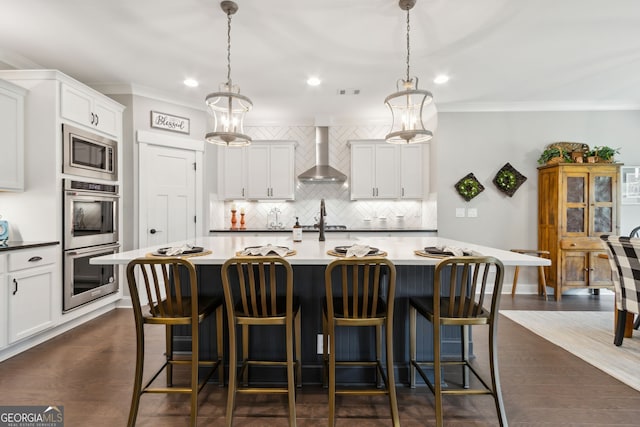 kitchen with wall chimney range hood, hanging light fixtures, appliances with stainless steel finishes, and a large island with sink