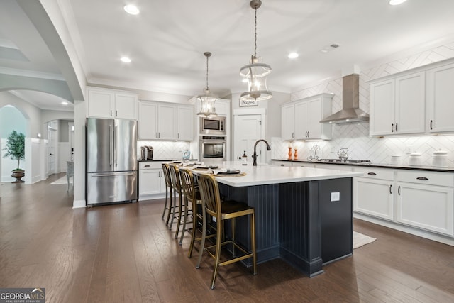 kitchen with wall chimney range hood, appliances with stainless steel finishes, an island with sink, dark hardwood / wood-style flooring, and decorative light fixtures