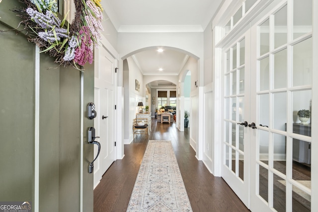 hall with a decorative wall, crown molding, arched walkways, french doors, and dark wood-type flooring