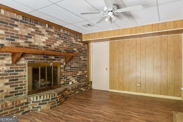 unfurnished living room with dark hardwood / wood-style floors, wooden walls, a drop ceiling, a brick fireplace, and ceiling fan