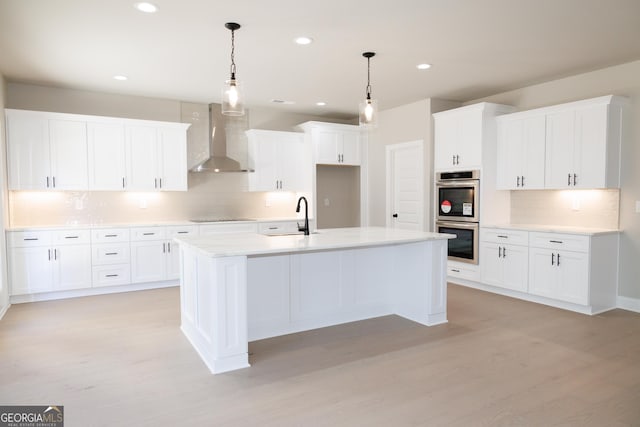 kitchen with wall chimney exhaust hood, stainless steel double oven, decorative light fixtures, light hardwood / wood-style floors, and an island with sink