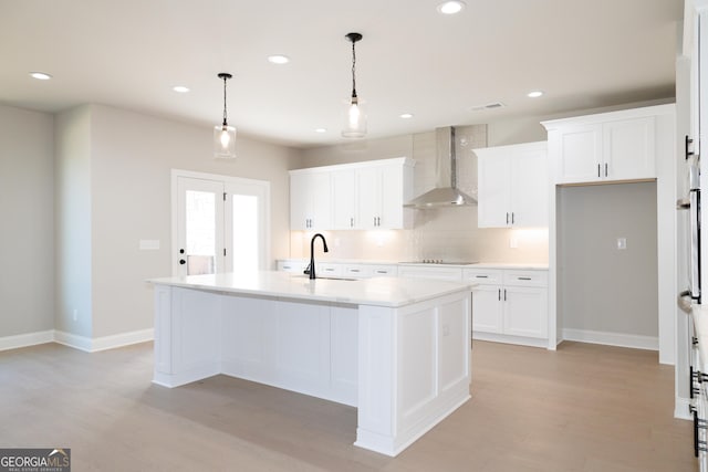 kitchen with wall chimney exhaust hood, a kitchen island with sink, sink, white cabinetry, and hanging light fixtures