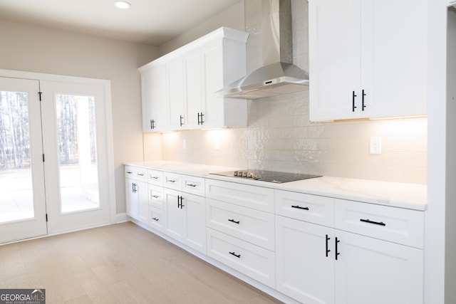 kitchen featuring black electric stovetop, backsplash, wall chimney range hood, white cabinets, and light hardwood / wood-style floors