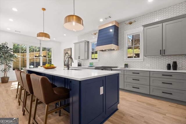 kitchen with gray cabinetry, sink, an island with sink, decorative light fixtures, and custom exhaust hood