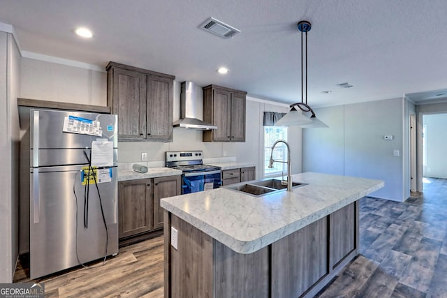 kitchen featuring wall chimney exhaust hood, sink, appliances with stainless steel finishes, pendant lighting, and a kitchen island with sink