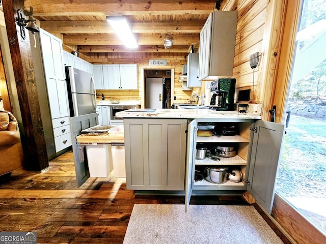 kitchen with stainless steel refrigerator, beamed ceiling, white cabinets, wooden walls, and dark hardwood / wood-style flooring