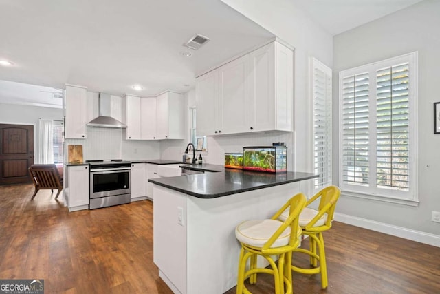 kitchen featuring a breakfast bar area, kitchen peninsula, wall chimney exhaust hood, white cabinets, and stainless steel range with electric cooktop