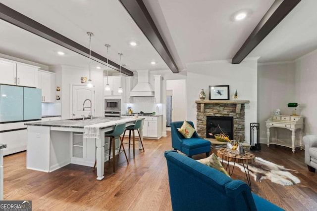 kitchen featuring white cabinets, premium range hood, a kitchen island with sink, beamed ceiling, and decorative light fixtures