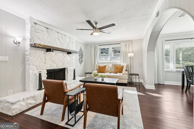 living room featuring a stone fireplace, a textured ceiling, ceiling fan, dark wood-type flooring, and ornamental molding