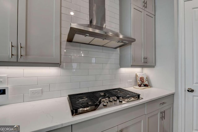 kitchen featuring tasteful backsplash, wall chimney exhaust hood, gray cabinets, stainless steel gas stovetop, and light stone counters