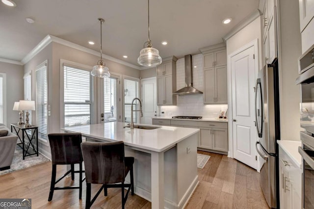 kitchen with wall chimney range hood, sink, stainless steel appliances, light hardwood / wood-style flooring, and a kitchen island with sink